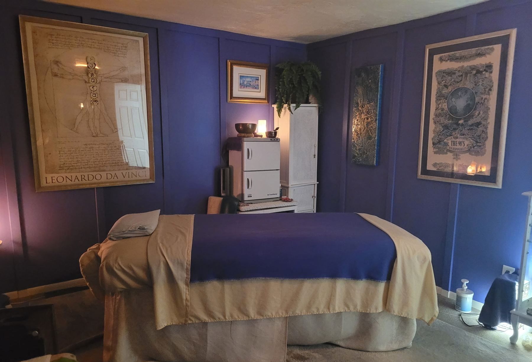 Stephanie&rsquo;s treatment room: a dressed massage table under warm lamplight against deep violet walls, framed prints and candles on either side.